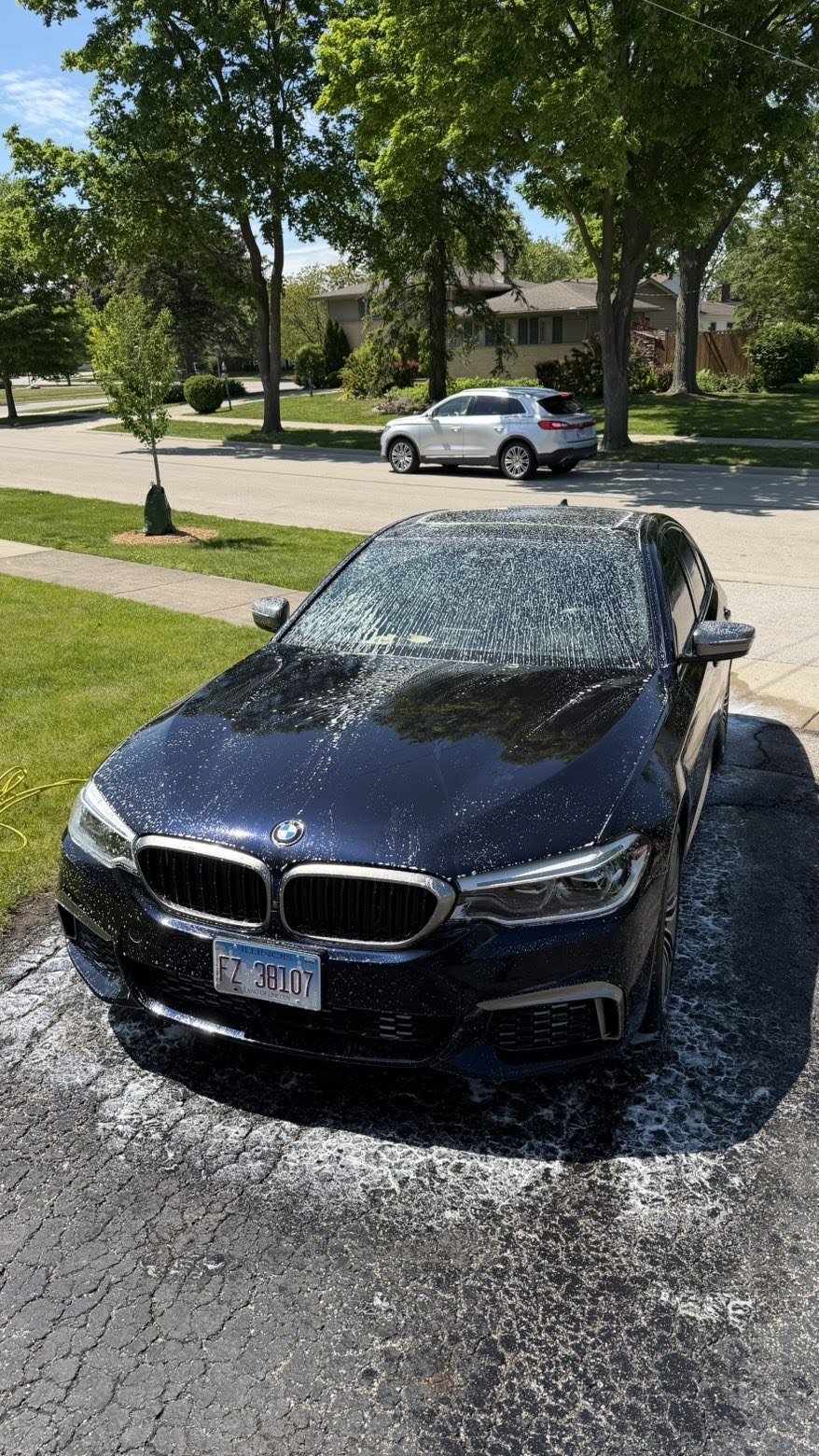 Dark blue BMW sedan parked in driveway with water droplets on hood, residential street and homes visible in background