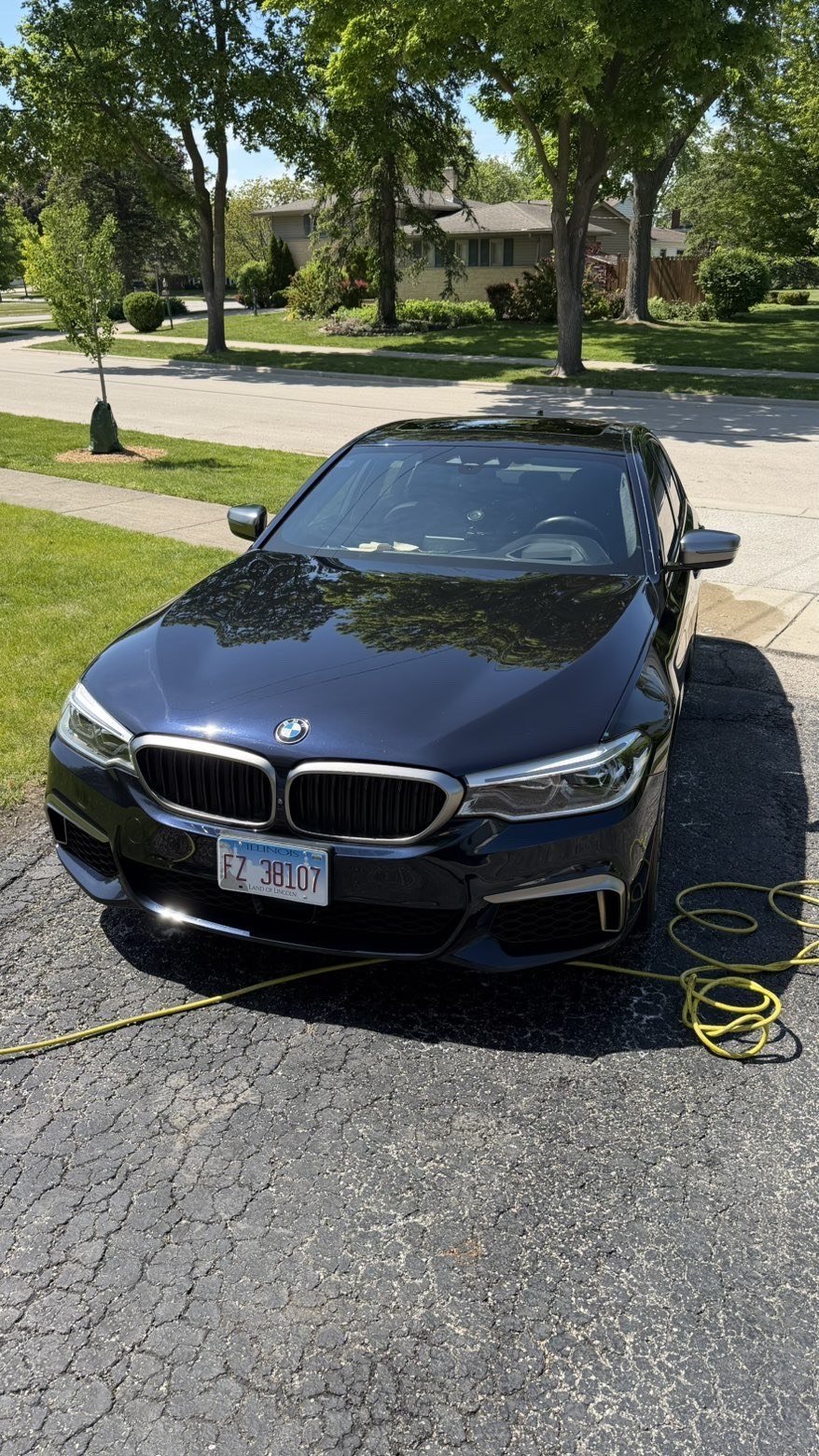 Black BMW sedan parked in a residential driveway with a yellow charging cable, tree-lined street and brick house visible in background